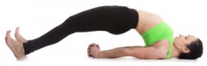 A woman in yoga gear lying on her back, legs and torso arched upward, arms under the body, hands clasped