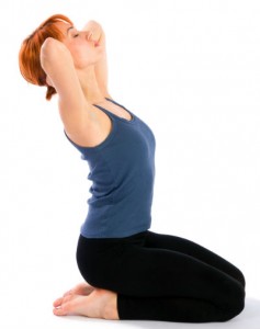 A woman in yoga gear on her knees, torso upright and stretching back a little, hands laced behind her head