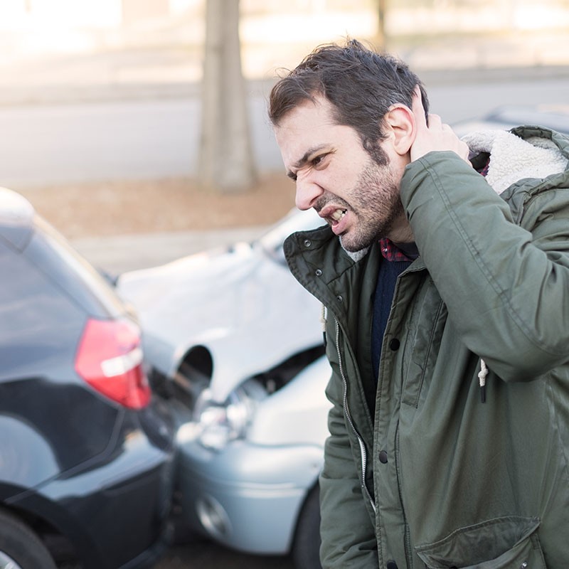 Man with Whiplash from Car Accident Man with whiplash cringing in pain, 2 smashed cars in the background